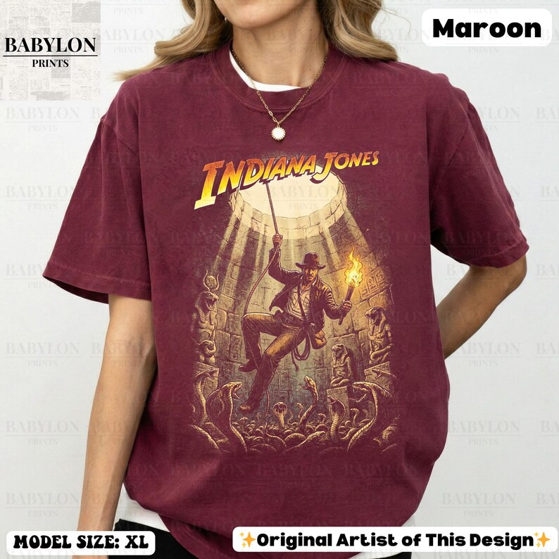 May include: Maroon t-shirt featuring the Indiana Jones movie poster graphic. The design shows Indiana Jones in a temple with snakes. The shirt has the text "INDIANA JONES" in yellow. The shirt is a size XL.