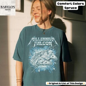 May include: Spruce-colored t-shirt featuring a graphic of the Millennium Falcon with the text "MILLENNIUM FALCON" in a metallic font. The design includes lightning bolts and the words "Comfort Colors" and "Original Artist of This Design".