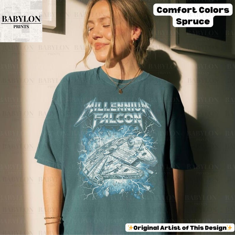 May include: Spruce-colored t-shirt featuring a graphic of the Millennium Falcon with the text "MILLENNIUM FALCON" in a metallic font. The design includes lightning bolts and the words "Comfort Colors" and "Original Artist of This Design".