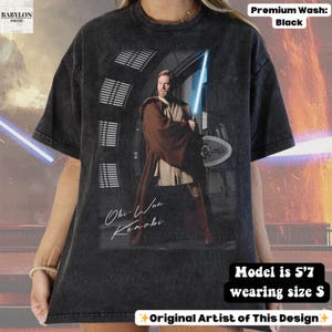 Może przedstawiać: Czarny t-shirt z grafiką Obi-Wana Kenobiego trzymającego miecz świetlny. Na koszulce napisano "Obi-Wan Kenobi" kursywą. Koszulka jest wysokiej jakości, a modelka ma 170 cm wzrostu i nosi rozmiar S.
