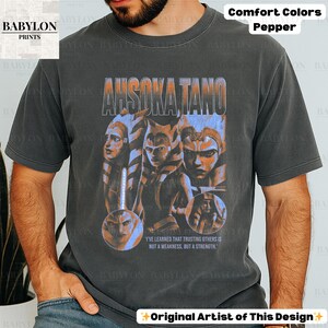 May include: A black t-shirt with a faded vintage look featuring a graphic of Ahsoka Tano from Star Wars. The graphic includes a quote from the show: "I've learned that trusting others is not a weakness, but a strength."