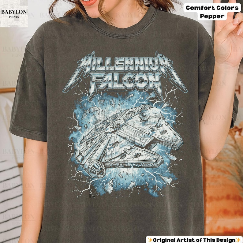 May include: Dark grey t-shirt featuring a graphic of the Millennium Falcon spaceship with the text "Millennium Falcon" in a stylized font. The design includes blue lightning and a distressed effect. The shirt is labeled "Comfort Colors Pepper".