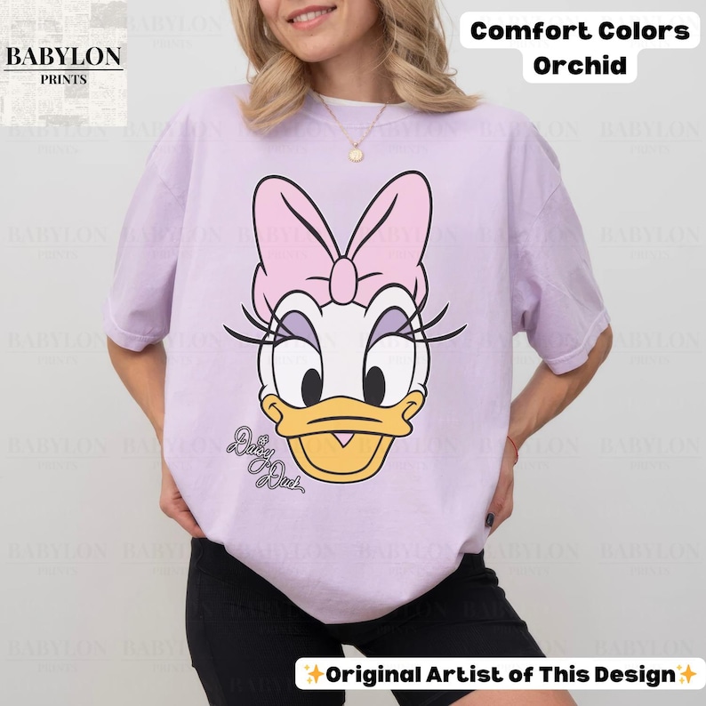 May include: A light purple Comfort Colors t-shirt featuring a Daisy Duck graphic. The graphic includes a pink bow, eyelashes, and the text "Daisy Duck". The shirt also has the text "Comfort Colors Orchid" and "Original Artist of This Design".