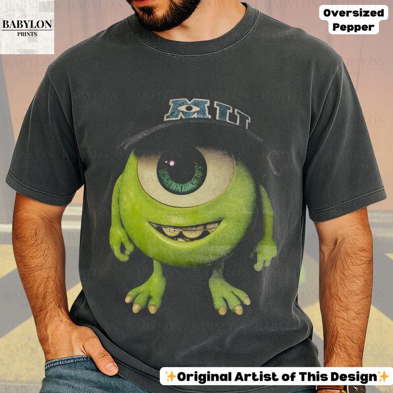 K&ouml;nnte beinhalten: Dunkelgraues T-Shirt mit einer gro&szlig;en Grafik von Mike Wazowski aus Monsters, Inc. Die Figur ist gr&uuml;n mit einem gro&szlig;en Auge und einem Hut mit der Aufschrift "MIT". Das Shirt ist mit "Oversized Pepper" und "Original Artist of This Design" beschriftet.