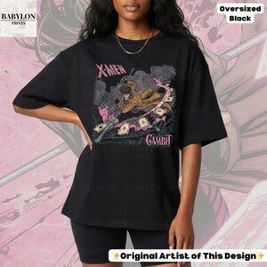 May include: Black oversized t-shirt featuring a graphic of the X-Men character Gambit. The design includes the text "X-Men" and "Gambit" in pink, along with an illustration of the character. The shirt is labeled "Oversized Black" and "Original Artist of This Design."