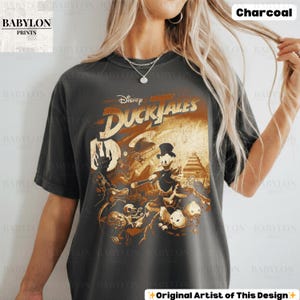 May include: Charcoal-colored t-shirt featuring a vintage-style graphic from Disney's DuckTales. The design includes Scrooge McDuck and other characters in a sepia-toned illustration. The shirt has the text "Disney DuckTales" across the top.