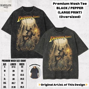 May include: Two black oversized t-shirts with a distressed graphic of Indiana Jones. The shirts feature the text "Indiana Jones" in yellow and a size chart. The shirts are labeled "Premium Wash Tee".
