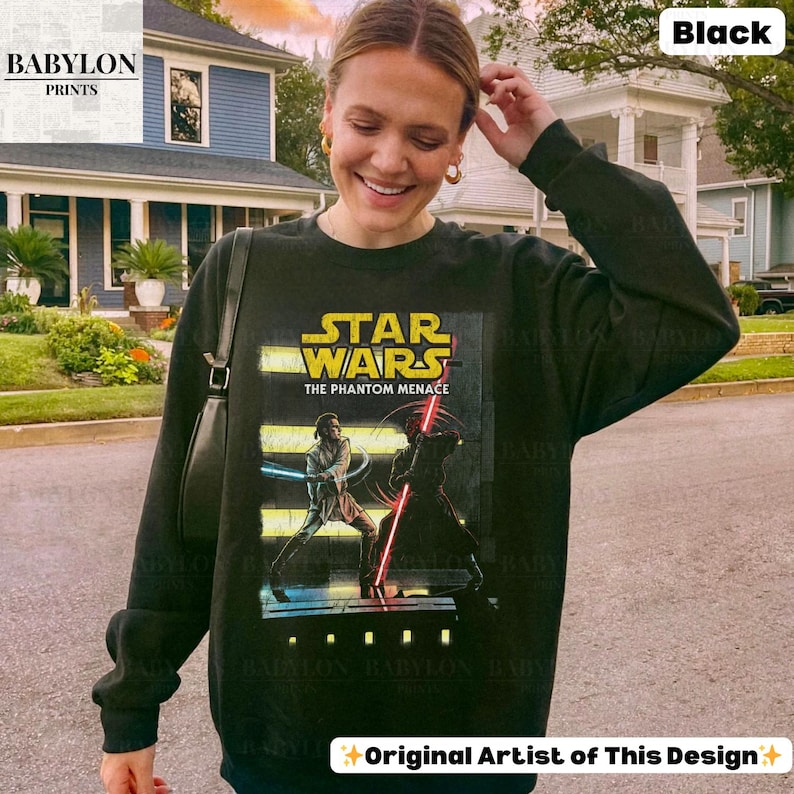 Puede incluir: Sudadera negra de Star Wars: La Amenaza Fantasma. El gr&aacute;fico presenta una escena con dos personajes empu&ntilde;ando sables de luz. La sudadera tiene el texto "STAR WARS THE PHANTOM MENACE" en amarillo y blanco. El logotipo de Babylon Prints est&aacute; en la esquina superior izquierda.