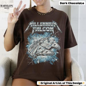 May include: Dark chocolate-colored t-shirt featuring a graphic of the Millennium Falcon spaceship with the text "Millennium Falcon" in a stylized font. The design includes lightning bolts and a distressed effect, creating a vintage look. The shirt is a relaxed fit.