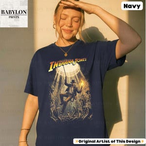 May include: Navy blue t-shirt featuring the text "Indiana Jones" in yellow and orange. The graphic depicts Indiana Jones in a cave with a torch and a whip. The shirt is a classic crew neck.