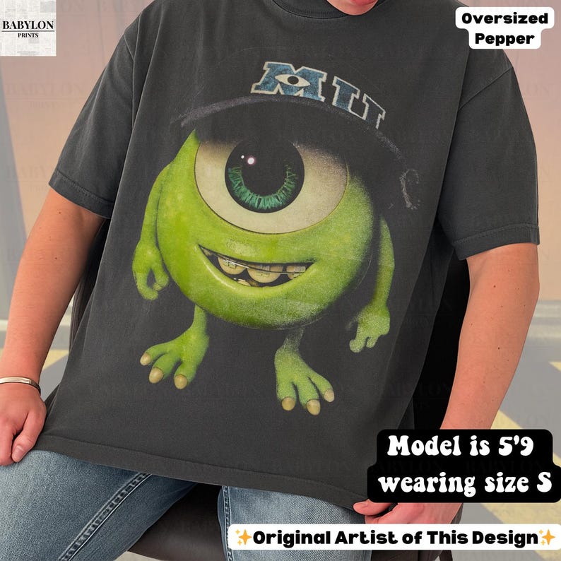 K&ouml;nnte beinhalten: Dunkelgraues, &uuml;bergro&szlig;es T-Shirt mit einer gro&szlig;en Grafik von Mike Wazowski aus Monsters, Inc., der einen schwarzen Hut mit "MIT" tr&auml;gt. Das Design ist in Gr&uuml;n, Schwarz und Wei&szlig; gehalten. Das T-Shirt hat kurze &Auml;rmel und einen Rundhalsausschnitt.