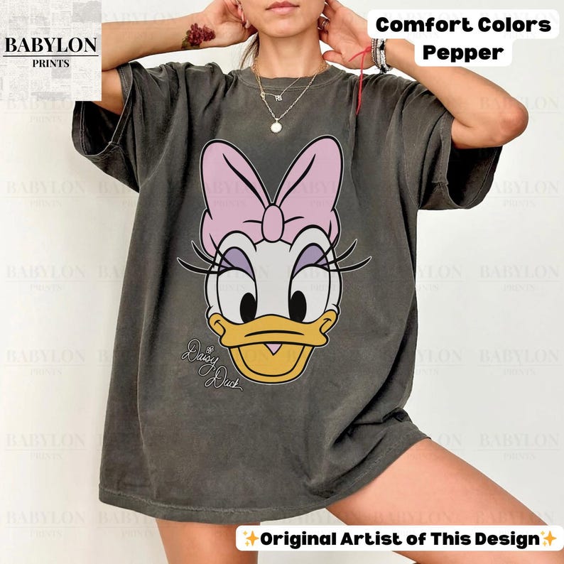 May include: A dark gray Comfort Colors t-shirt featuring a large graphic of Daisy Duck with a pink bow. The text "Daisy Duck" is written in cursive below the character's face. The shirt also has the text "Comfort Colors Pepper" and "Original Artist of This Design".