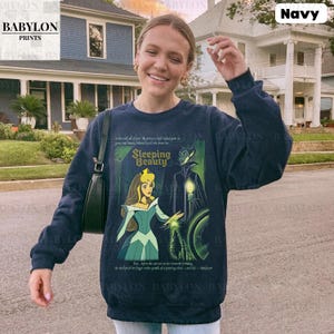 May include: Navy blue sweatshirt featuring a graphic from the Disney movie "Sleeping Beauty." The design includes Princess Aurora and Maleficent. The sweatshirt has long sleeves and a crew neck. The text "Sleeping Beauty" is displayed prominently.
