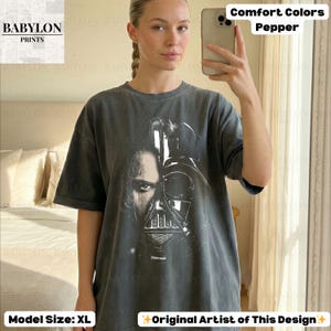 May include: Dark grey t-shirt featuring a split image of Darth Vader and Anakin Skywalker. The shirt has the text "Comfort Colors Pepper" and "Original Artist of This Design". The model is wearing a size XL.