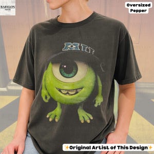 K&ouml;nnte beinhalten: Dunkelgraues, &uuml;bergro&szlig;es T-Shirt mit einer gr&uuml;nen Cartoonfigur mit einem Auge und einem Hut mit der Aufschrift "MIT". Das Shirt tr&auml;gt auch den Text "Oversized Pepper" und "Original Artist of This Design".