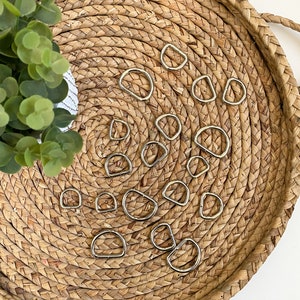May include: A collection of silver D-rings of various sizes arranged on a woven straw tray. The D-rings are commonly used for crafting, leatherwork, and jewelry making.