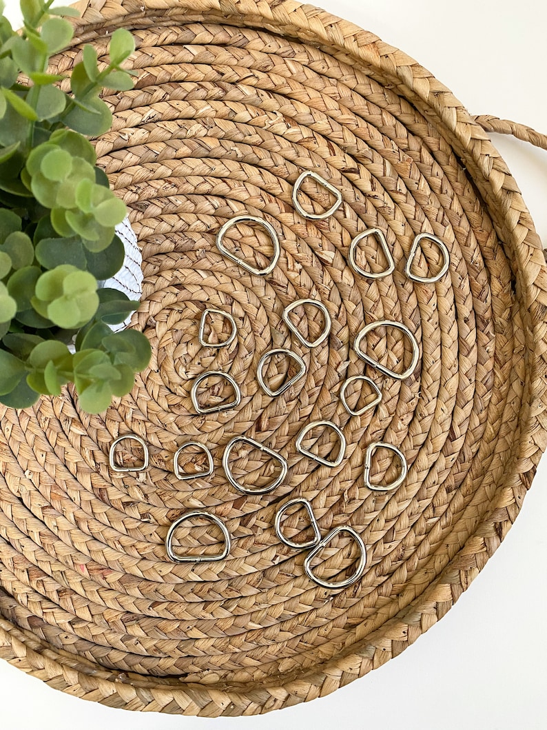 May include: A collection of silver D-rings of various sizes arranged on a woven straw tray. The D-rings are commonly used for crafting, leatherwork, and jewelry making.