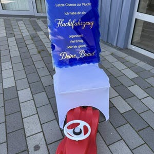 Könnte beinhalten: Ein rotes Spielzeugauto steht unter einem Stuhl, der für eine Hochzeit dekoriert ist. Der Stuhl hat eine weiße Hülle und ein Schild mit deutschem Text in Blau und Gold, einschließlich "Fluchtfahrzeug" (Fluchtfahrzeug).