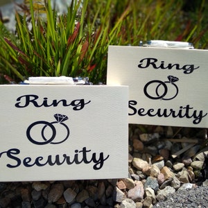 Könnte beinhalten: Zwei weiße Holzkisten mit dem Text "Ring Security" und einer Grafik von zwei Ringen mit einem Diamanten oben drauf.