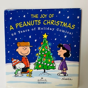 May include: A square book cover with a blue background and white stars. The title "The Joy of a Peanuts Christmas" is in yellow. Charlie Brown, Snoopy, Lucy, and a Christmas tree are featured. The Hallmark Books logo is at the bottom.