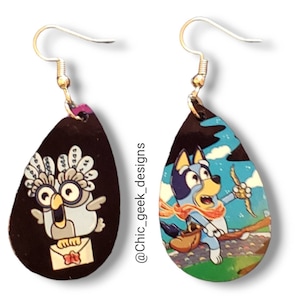 May include: Teardrop-shaped earrings featuring cartoon characters. One earring depicts a white owl holding a letter on a black background. The other earring shows a blue dog with a scarf and a bow and arrow against a sky background.