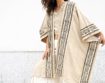 May include: A woman wearing a beige kimono with black and brown geometric embroidery. The kimono has long sleeves and a relaxed fit. The woman is smiling and has curly brown hair.