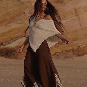 May include: A cream-colored, loose-fitting, short-sleeved top paired with wide-leg, dark brown trousers. The trousers feature a wide, cream-colored hem with a detailed, repeating geometric pattern and a frayed edge along the bottom hemline.