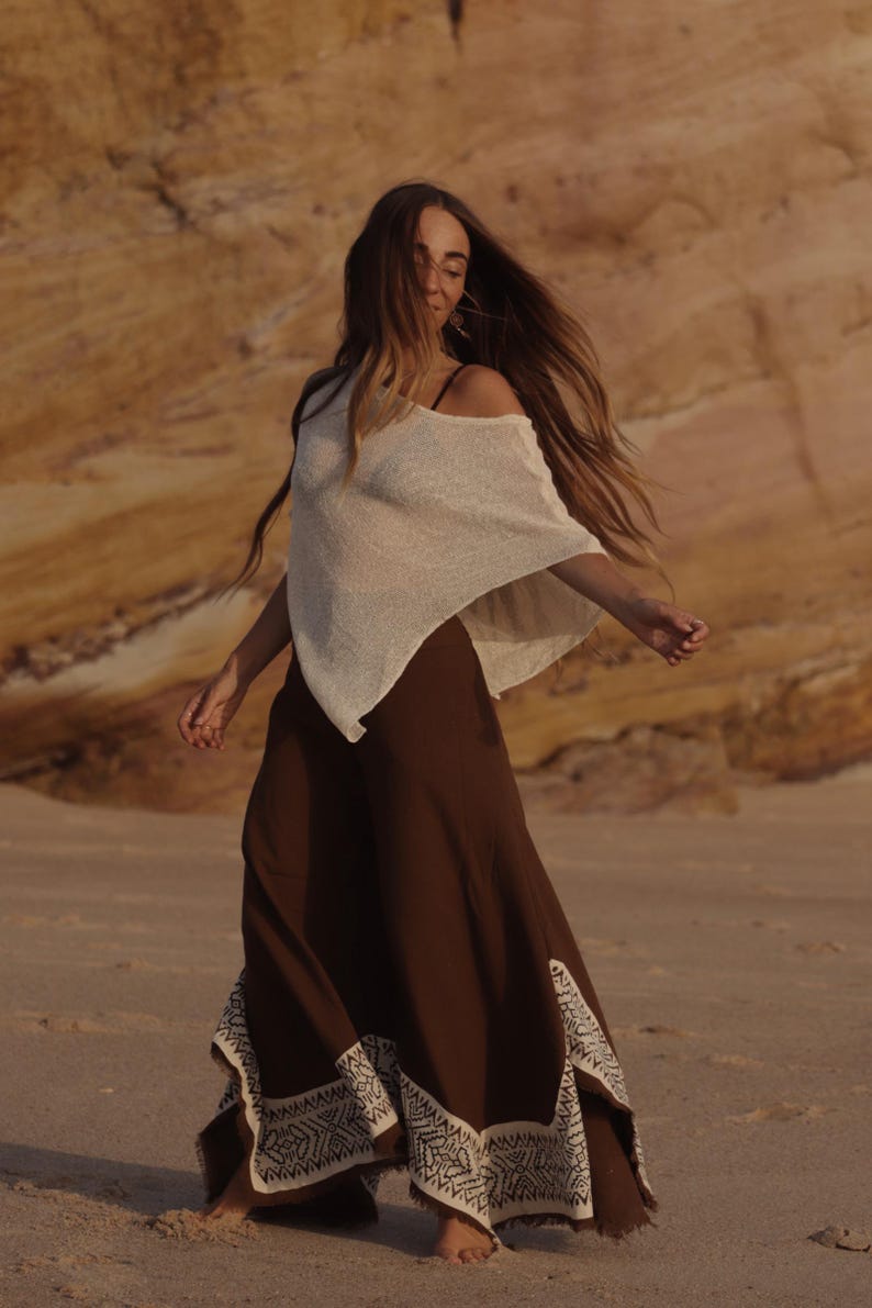 May include: A cream-colored, loose-fitting, short-sleeved top paired with wide-leg, dark brown trousers. The trousers feature a wide, cream-colored hem with a detailed, repeating geometric pattern and a frayed edge along the bottom hemline.