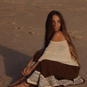 May include: A long-sleeved off-the-shoulder cream-colored top paired with a dark brown skirt featuring a wide white hem decorated with a black geometric pattern. The skirt has a fringed edge along the bottom and is worn with a beaded anklet.