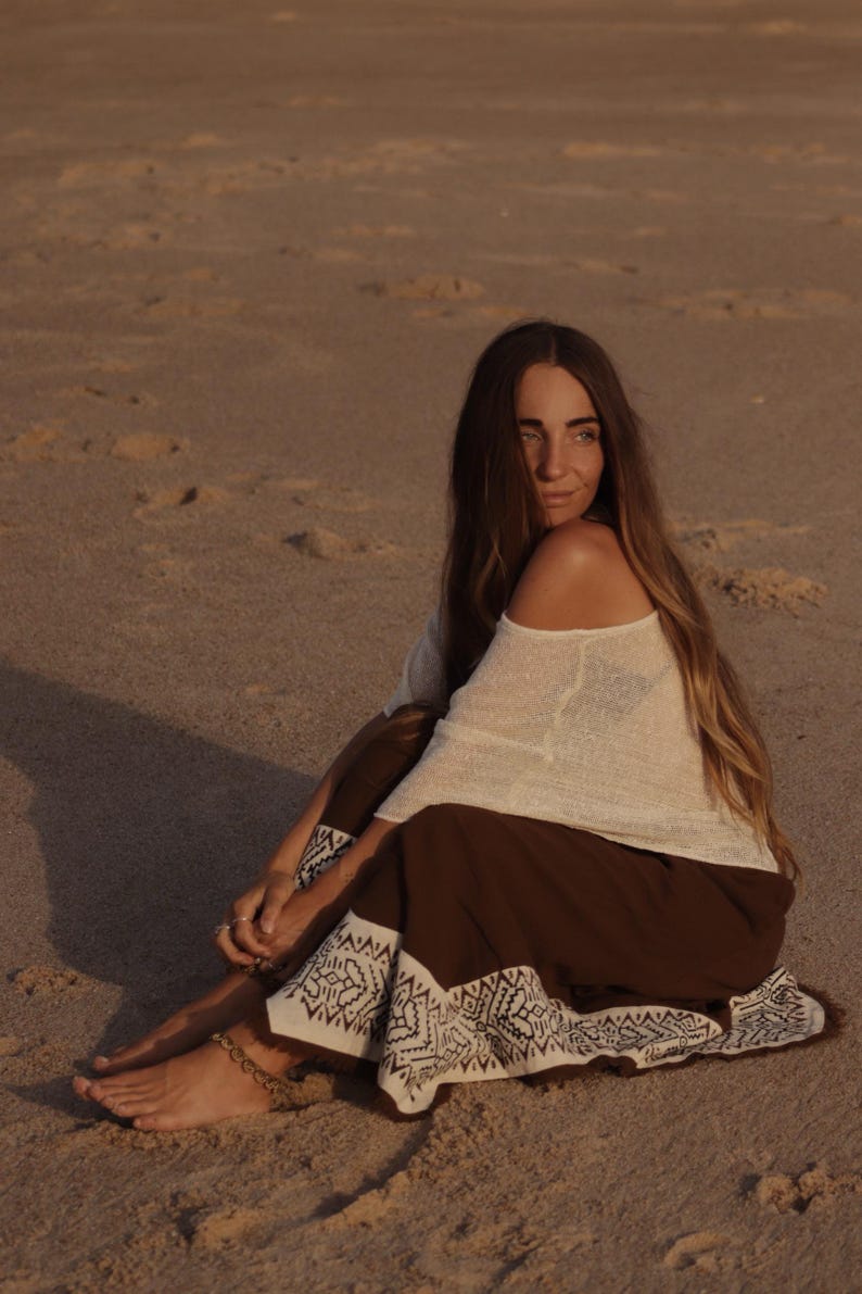May include: A long-sleeved off-the-shoulder cream-colored top paired with a dark brown skirt featuring a wide white hem decorated with a black geometric pattern. The skirt has a fringed edge along the bottom and is worn with a beaded anklet.
