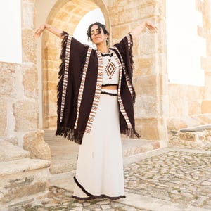 Peut inclure: Une femme portant un poncho brun à franges et une jupe blanche à franges. Elle se tient dans une arche de pierre.