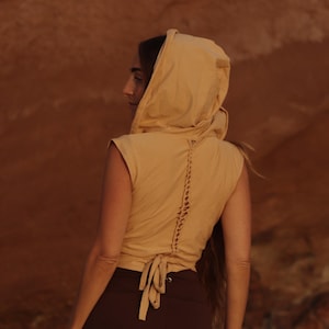 Peut inclure: Un haut à capuche sans manches de couleur jaune moutarde, doté d'un laçage au dos avec des liens en tissu à la taille. Le vêtement présente une capuche et une coupe ajustée, avec une fermeture à cordon verticale le long du milieu du dos.