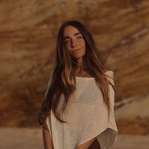 May include: A cream-colored, loose-fitting, short-sleeved knit top with a wide neckline and a semi-sheer, textured weave. The garment features a relaxed, boxy silhouette that drapes over the shoulders and torso, paired with dark brown trousers.