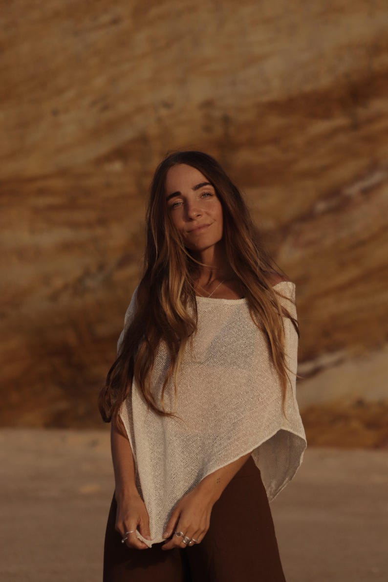 May include: A cream-colored, loose-fitting, short-sleeved knit top with a wide neckline and a semi-sheer, textured weave. The garment features a relaxed, boxy silhouette that drapes over the shoulders and torso, paired with dark brown trousers.