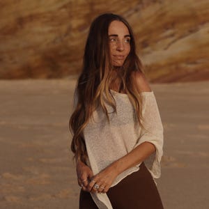 May include: A loose-fitting, off-the-shoulder cream-colored knit top with a semi-sheer texture and wide sleeves, paired with solid dark brown trousers. The top features a wide, relaxed neckline and a lightweight, breathable weave construction.