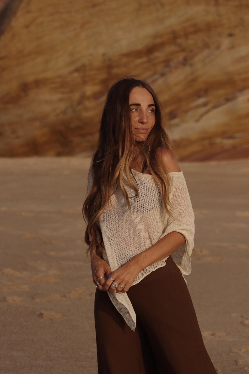 May include: A loose-fitting, off-the-shoulder cream-colored knit top with a semi-sheer texture and wide sleeves, paired with solid dark brown trousers. The top features a wide, relaxed neckline and a lightweight, breathable weave construction.
