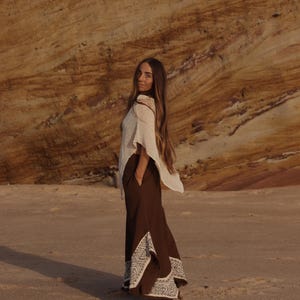 May include: A cream-colored sleeveless top paired with dark brown wide-leg trousers featuring white embroidered geometric patterns along the lower hem and side slits. The trousers include side pockets and a loose, flowing silhouette.