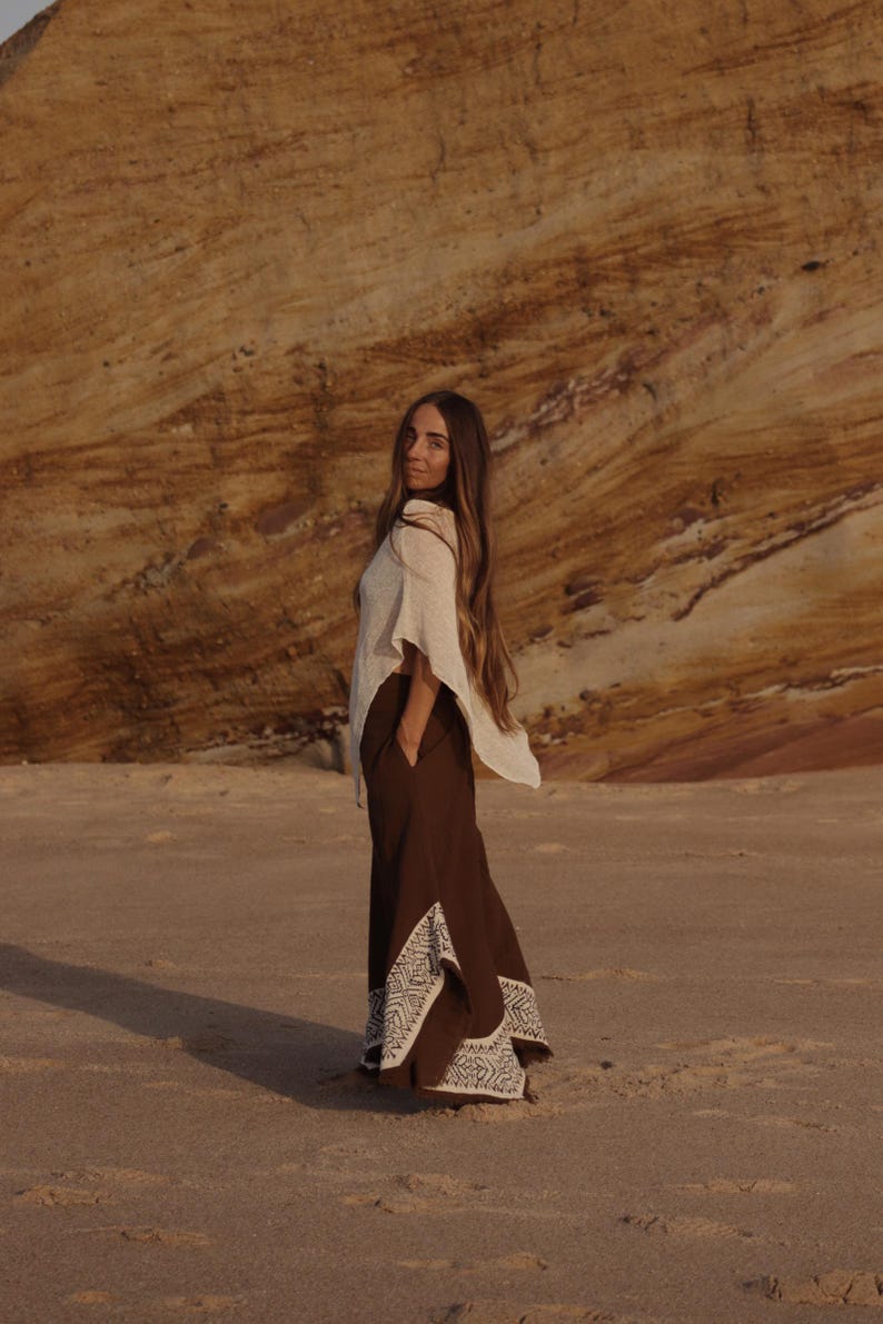 May include: A cream-colored sleeveless top paired with dark brown wide-leg trousers featuring white embroidered geometric patterns along the lower hem and side slits. The trousers include side pockets and a loose, flowing silhouette.