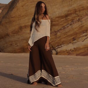 May include: A cream-colored, off-the-shoulder, lightweight knit top paired with wide-leg, dark brown trousers. The trousers feature a wide, cream-colored hem band decorated with a repeating geometric pattern in a dark brown, intricate design.
