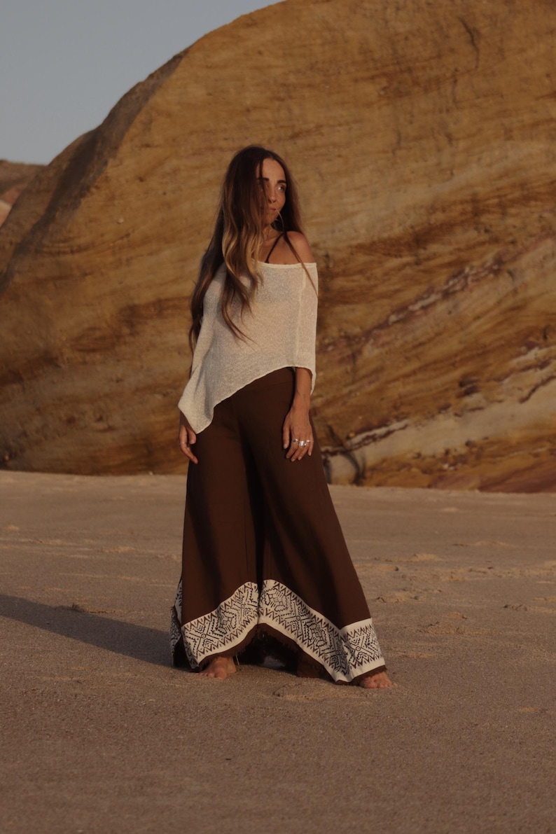 May include: A cream-colored, off-the-shoulder, lightweight knit top paired with wide-leg, dark brown trousers. The trousers feature a wide, cream-colored hem band decorated with a repeating geometric pattern in a dark brown, intricate design.