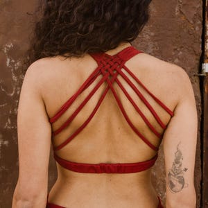 May include: A red crop top with a distinctive woven back. The top has multiple straps that cross and weave together, forming an intricate pattern. The garment is made of a soft, flowing fabric. The model is wearing a matching red skirt.