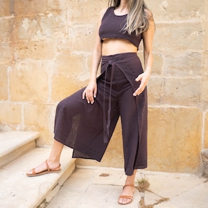 May include: A woman wearing a brown crop top and matching wide-leg pants with a wrap-around design. She is standing on a set of stone steps with a stone wall in the background.