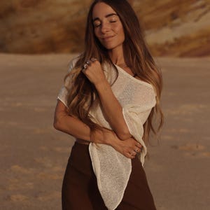 May include: A cream-colored, short-sleeved, loose-knit top with a wide neckline and a pointed hemline, paired with solid dark brown high-waisted trousers. The top features a semi-sheer, textured weave, while the trousers have a smooth, matte finish.