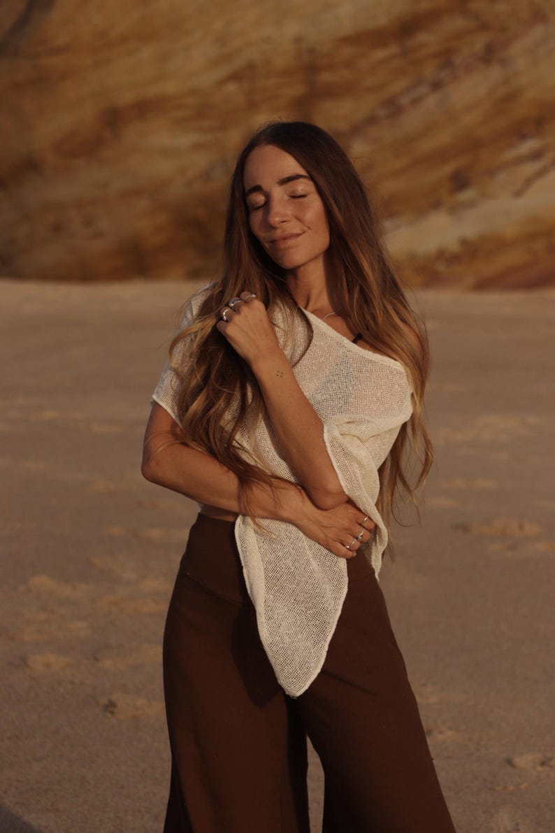 May include: A cream-colored, short-sleeved, loose-knit top with a wide neckline and a pointed hemline, paired with solid dark brown high-waisted trousers. The top features a semi-sheer, textured weave, while the trousers have a smooth, matte finish.