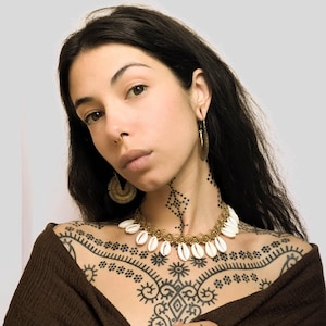 Può includere: Una donna con lunghi capelli scuri indossa un maglione marrone e una collana d'oro con conchiglie bianche. Ha un tatuaggio geometrico sul petto e sulle braccia.