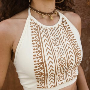 May include: Cream halter top with a brown geometric design. The top has a halter neck and a fitted shape. The brown pattern includes arrows and other shapes. The top is likely made of a soft, breathable fabric.