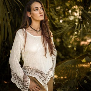 Może przedstawiać: Kremowy top z długim rękawem, okrągłym dekoltem i dekoracyjnym brązowym wzorem wzdłuż dołu i rękawów. Top jest połączony z brązowymi spodniami. Zdjęcie zrobione na zewnątrz z naturalnym tłem.