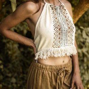 Puede incluir: Top halter crema con patrones geométricos marrones y detalles de flecos, combinado con pantalones marrones con cordón. El top presenta un cuello halter y un diseño decorativo en el pecho. Los pantalones son de color liso y de corte holgado.