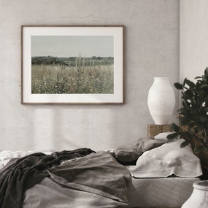 Puede incluir: Un interior de dormitorio con una cama con ropa de cama gris y negra, una fotograf&iacute;a de paisaje enmarcada en la pared, un jarr&oacute;n de cer&aacute;mica blanco sobre una mesa de madera y una planta en maceta en la esquina.