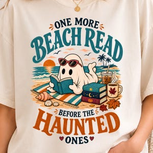 Pode incluir: Uma t-shirt cor de creme com um gráfico de um fantasma branco com óculos de sol vermelhos, lendo um livro numa toalha de praia. O texto diz ONE MORE BEACH READ BEFORE THE HAUNTED ONES. Inclui ilustrações de livros, café e conchas do mar.